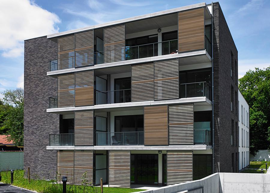 Modernes mehrstöckiges Wohnhaus mit dunkler Klinkerfassade und Holzlatten-Balkonverkleidung, blauer Himmel mit Wolken, gedämpfte grüne und braune Töne